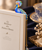 Marque-page paon bleu fleuri placé dans un livre avec longue chaînette dorée et breloque plume